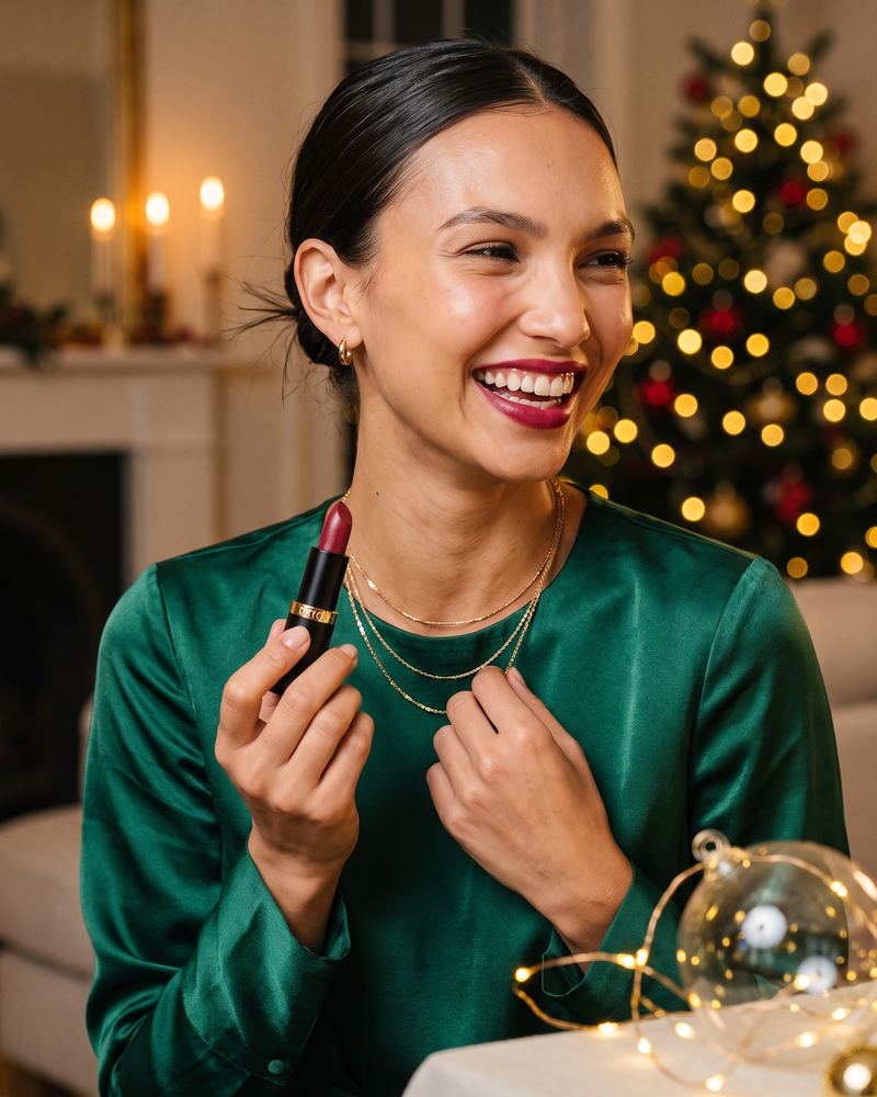Model laughing in green satin, holding a red lipstick by the Christmas tree